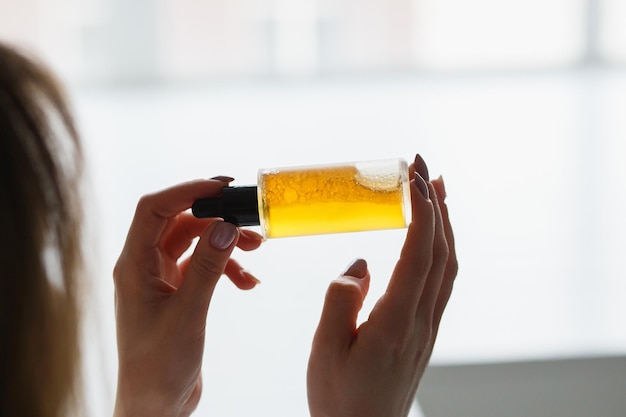 Hands of cropped white woman holding cosmetic serum bottle with pipette near window texture of serum