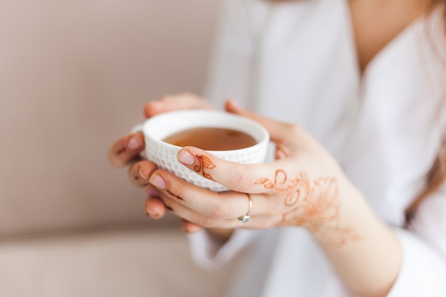 Photo in the hands of a cup of tea or coffee, a drink in the morning, lunch, hands covered with a traditional pattern, mahendi, drawing on the hands