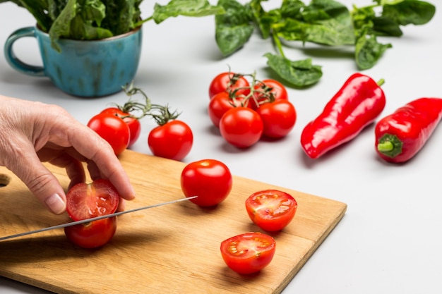 Hands cut red pepper with knife. Tomatoes and spinach leaves on table. Gray background. Top view.