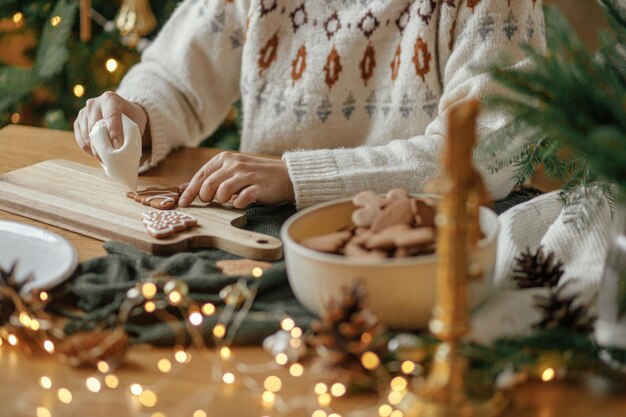 Фото Руки украшают рождественские имбирные печенье с глазурой на деревенском деревянном столе на фоне золотых огней атмосферные традиции рождественских праздников украшают печенье сахарной глазурой