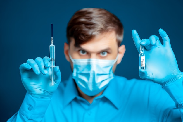 In the hands of a doctor, a doctor in a medical mask and gloves, dressed in a blue uniform, an injection syringe and a vaccine against coronavirus