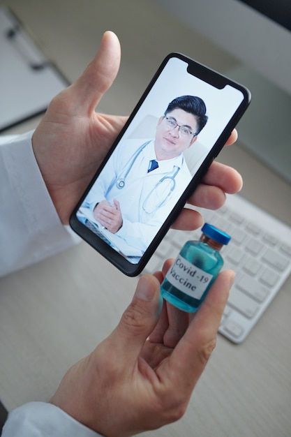 Hands of doctor holding vial of vaccine against coronavirus when video calling his colleague from another hospital