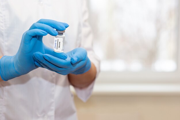 The hands of a doctor or nurse in blue gloves and a white coat are carefully holding a bottle of coronavirus vaccine. Close up. Copy space