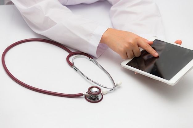 Hands of doctor in a white lab coat using tablet and putting stethoscope on a table.