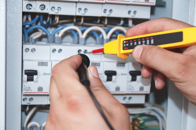 Hands of electrician with multimeter probe at an electrical switchgear cabinet examining fuse box