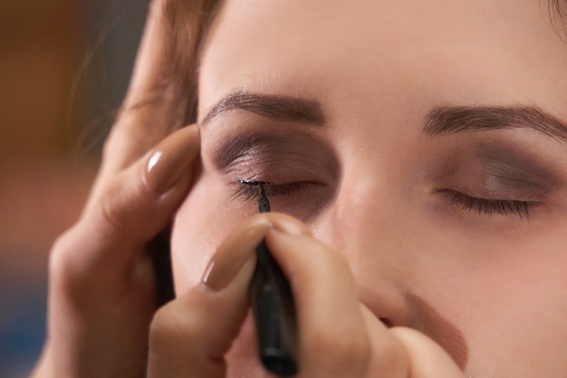 Hands of a female make-up artist draw eyeliner on the eyelids of a woman model, close-up