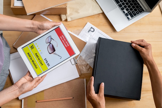 Photo hands of female managers of online shop holding tablet with photo of sunglasses on screen and packing black box with order of client