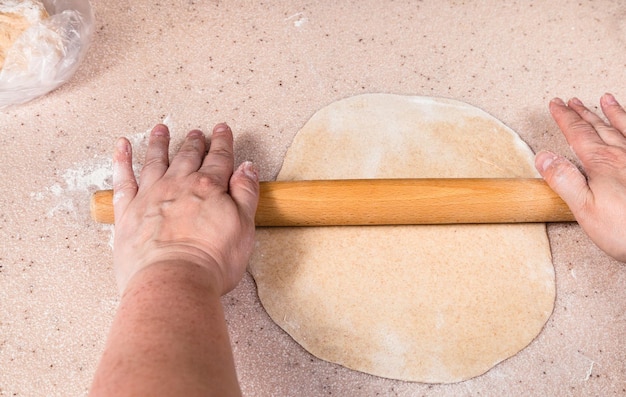 Hands flatten sheet of dough by wooden rolling pin