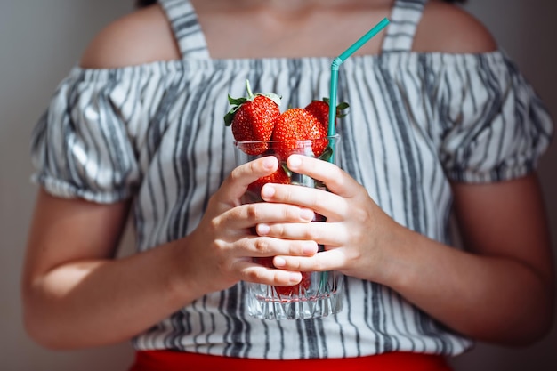 The hands of a girl in a striped blouse and a red skirt hold a glass with juicy strawberries