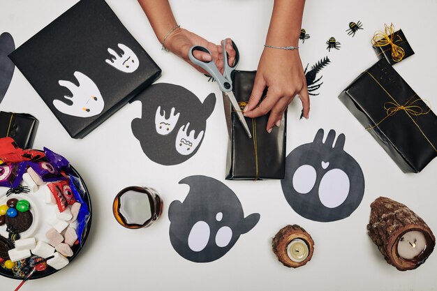 Hands of girl wrapping Halloween present in black paper and decorating with ghost stickers