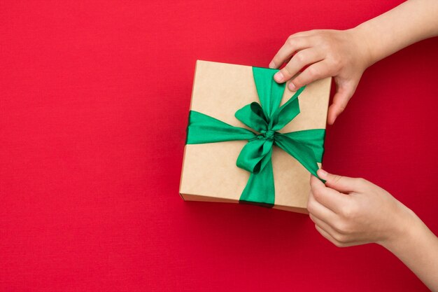 Руки дают или получают подарок из крафт-бумаги с зеленой лентой на красном. Вид сверху.