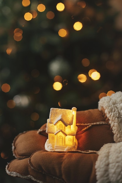 Hands in gloves holding little glowing house on background of illuminated christmas tree lights Magical winter time atmospheric image Cozy home Merry Christmas and Happy Holidays