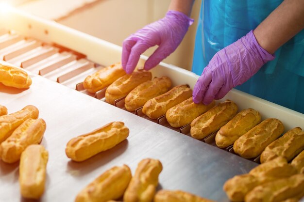 Hands in gloves touching eclairs Eclair shells on narrow conveyor Employee of confectionery plant Worker responsible for quality