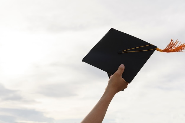 The hands of the graduates hold a black hat and a yellow tassel extends to the sky.