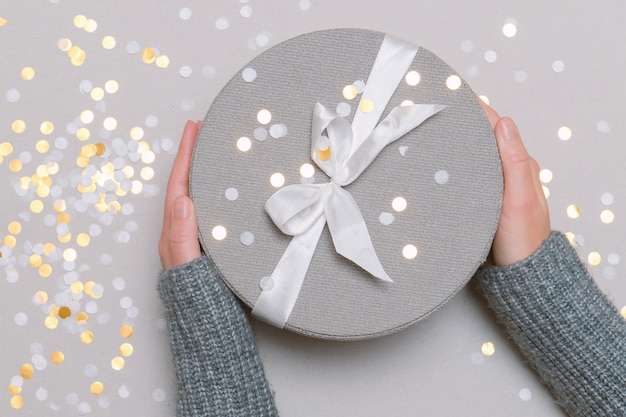 hands gray sweater holding round gift box with bow confetti top view