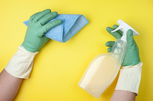 Hands in green gloves hold rag and detergents on yellow background. Cleaning or housekeeping concept.  Flat lay. Top view