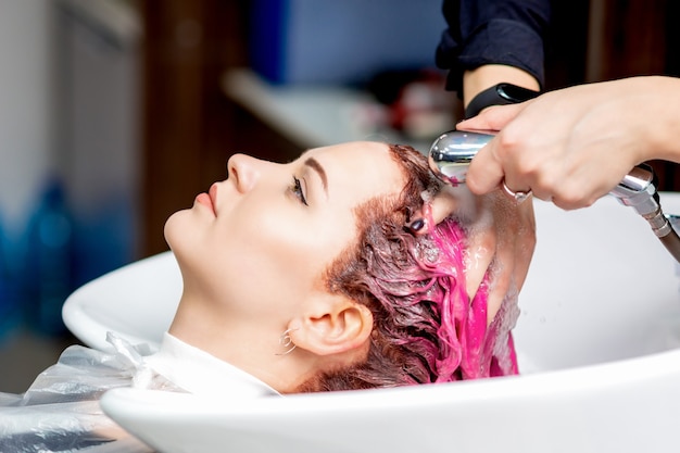 Hands of hairdresser are washing pink dyed hair of woman in sink at hair salon.