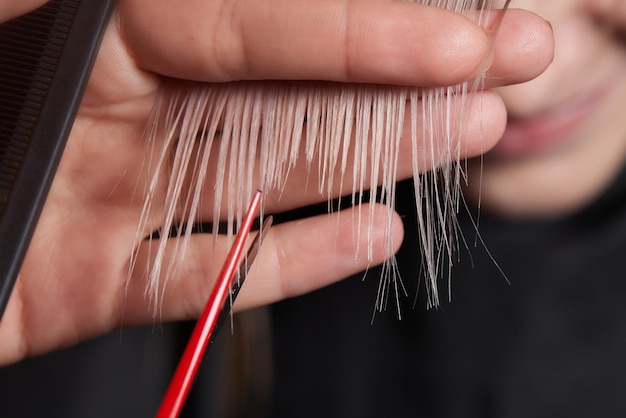 Le mani del parrucchiere tengono la ciocca di capelli tra le dita facendo il taglio di capelli di capelli lunghi della giovane donna con pettine e forbici nel salone di parrucchiere da vicino