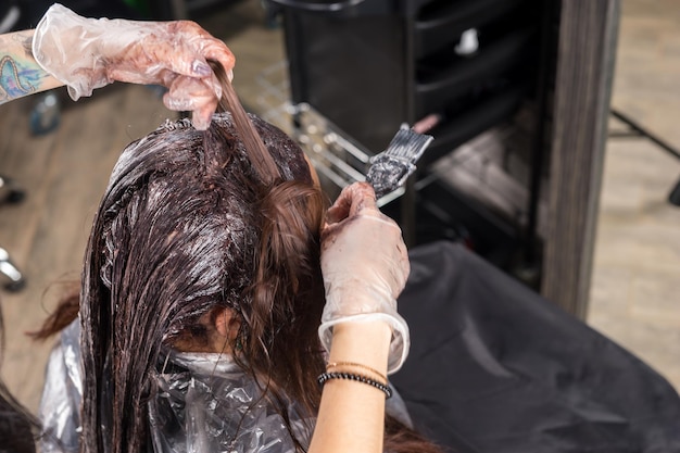 Hands of hairdresser thoroughly dyeing hair of female client using paintbrush while she is sitting in chair in beauty salon