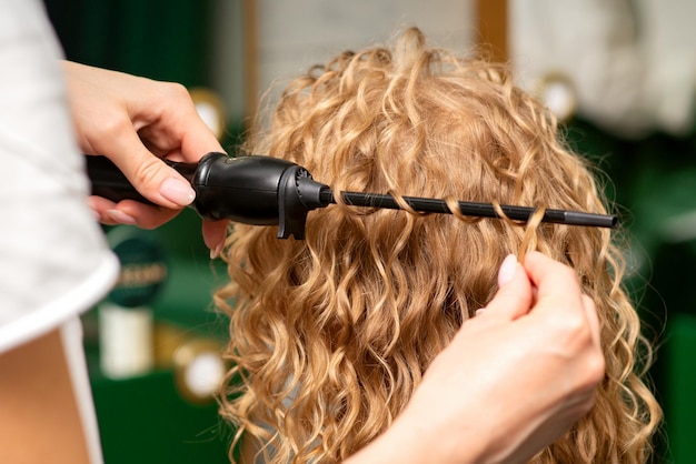 Hands of hairstylist curl wavy hair of young woman using a curling iron for hair curls in the beauty salon rear view.