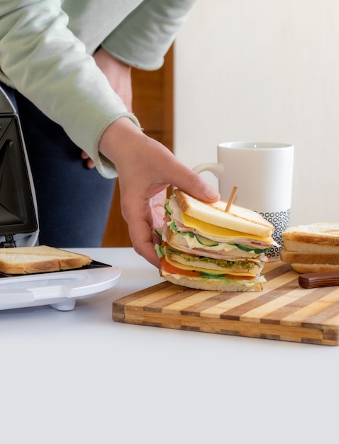 Hands hold cooked fresh sandwich with cheese, bacon and vegetable near toster and cup of coffee