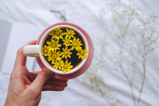 Hands hold cup with yellow flowers inside, on a white bed in the morning. Spring background