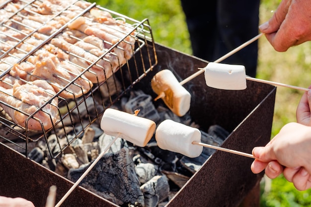 Hands hold marshmallows on a wooden skewer over the grill fried marshmallows and grilled meat