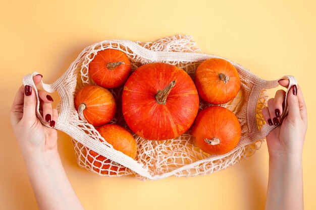 Hands hold mesh bag with ripe pumpkins on yellow background Autumn harvest and Thanksgiving concept