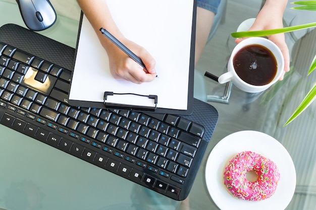 Photo hands hold a pen over a blank white of paper keyboard computer mouse donut on a plate and coffee