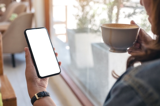 Photo hands holding black mobile phone with blank desktop screen while drinking coffee in cafe