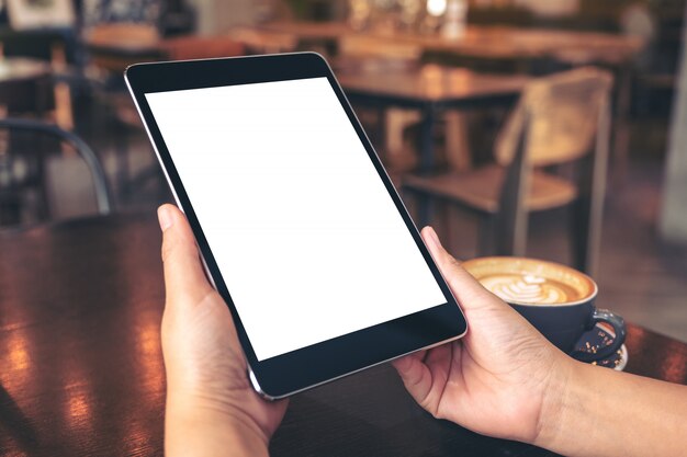  hands holding black tablet pc with blank white screen with coffee cup on wooden table in cafe