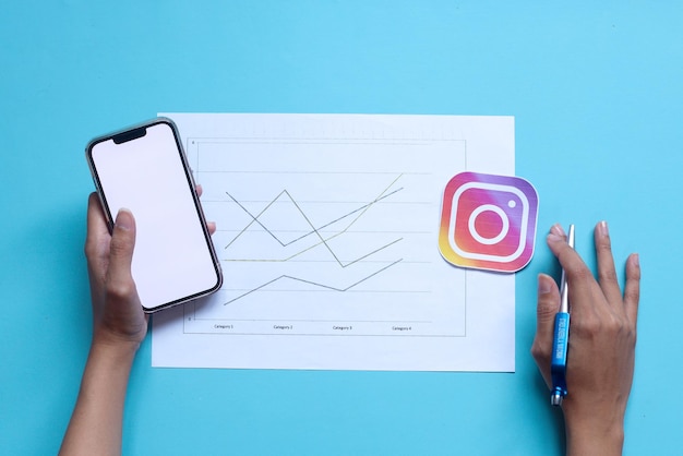 Hands holding blank screen of smartphone and pen with graph paper and Instagram logo icon. Instagram