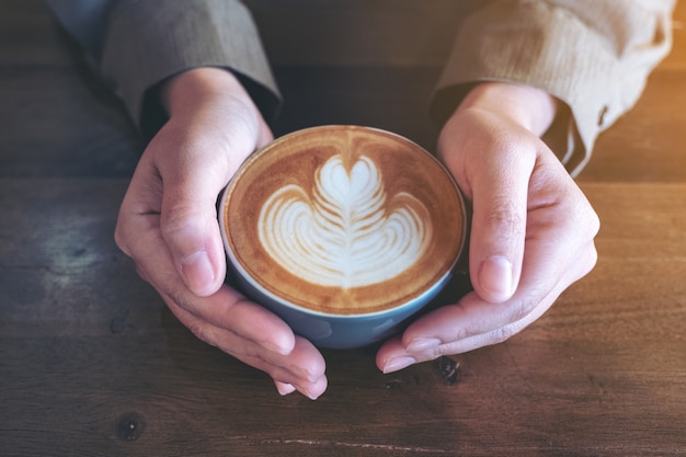 hands holding a blue cup of hot latte coffee with latte art on wooden table in cafe