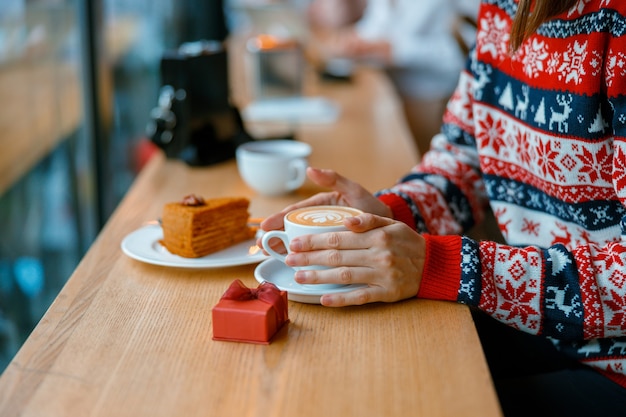 Hands holding a capuchino cup with gift box and cake on a cozy cafe terace