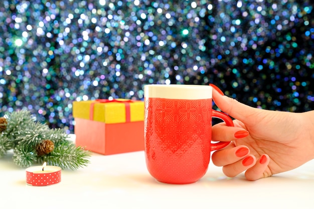 Hands holding a coffee mug on a Christmas background. View from above. Female hands holding coffee cup. Christmas gift boxes and snow fir tree above wooden table.