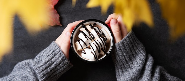 Hands holding a cup of hot chocolate with marshmallow on a dark\
gray background autumn concept of a warming drink top view\
horizontal orientation