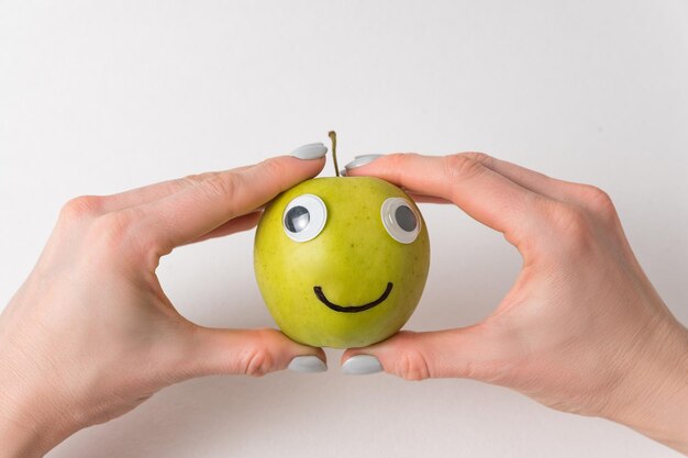 Hands holding cute little Apple with Googly eyes and drawn smile. Apple haracter on white background.