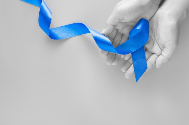Photo hands holding deep blue ribbon on blue background with copy space. colorectal cancer awareness colon cancer of older person and world diabetes day child abuse prevention. healthcare, insurance concept