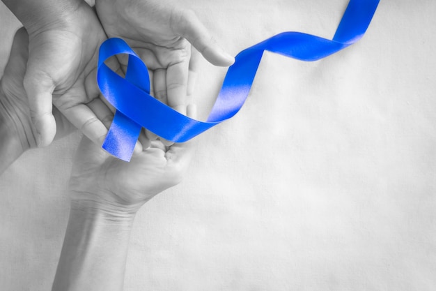 Photo hands holding deep blue ribbon on white fabric with copy space. colorectal cancer awareness, colon cancer of older person and world diabetes day, child abuse prevention. healthcare, insurance concept.