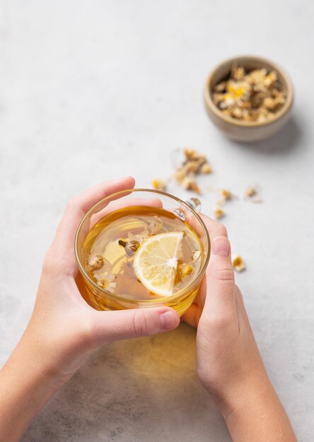 Hands holding a glass cup with chamomile herbal tea on a light background with dry flowers The concept of a healthy drink for health and immunity