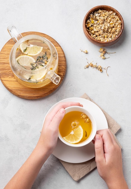 Руки, держащие стеклянную чашку с ромашковым травяным чаем на светлом фоне с сухими цветами и чайником Концепция здорового напитка для здоровья и иммунитета