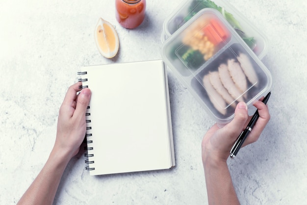 Le mani che tengono la scatola di pranzo sana e il taccuino in bianco per il vostro messaggio di testo o progettazione, ordinano l'alimento.