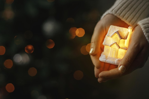 Hands holding illuminated little house on background of christmas tree with lights bokeh Cozy home Atmospheric Holiday banner Family and safety concept Merry Christmas