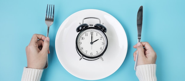 hands holding knife and fork above alarm clock on white plate