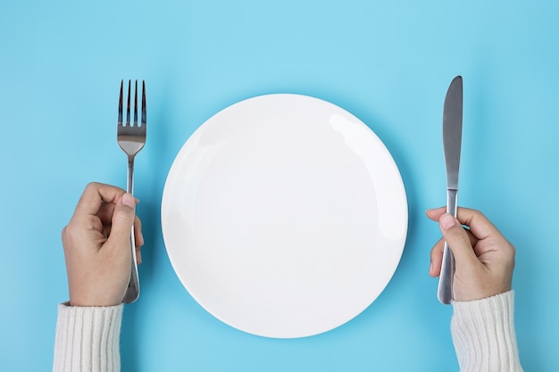Hands holding knife and fork above white plate on blue background., dieting, weight loss, dining and kitchenware concept