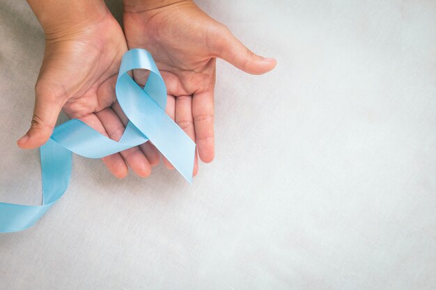 Hands holding light blue color ribbon on white fabric table with copy space Lymphedema and Prostate Cancer Awareness month symbol Healthcare medical and insurance concept Male cancer