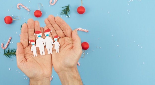 Hands holding a paper-cut family with masks at Christmas. New normality by coronavirus social distance at Christmas. Copy space.