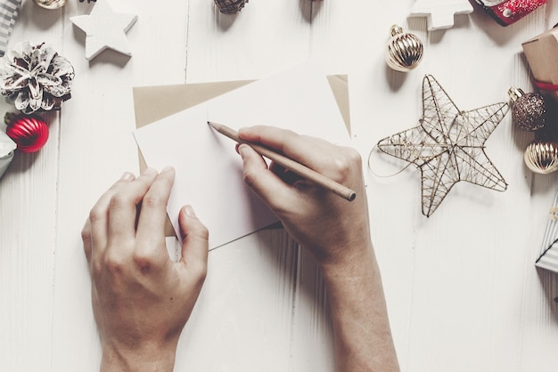 Hands holding pencil and writing a letter to santa claus with space for text merry christmas and happy new year craft and presents and ornaments on white wood person wish list for holidays