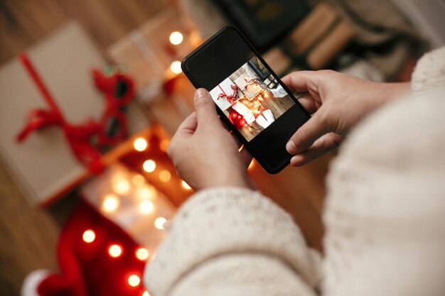 Руки держат телефон и фотографируют рождественские подарочные коробки, иллюминационные огни в шляпе Санты на деревянном фоне в темной комнате Стильная хипстерская девушка в свитере делает рождественское плоское фото