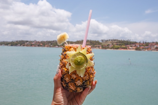 Mani che tengono bevanda all'ananas con sfondo spiaggia.